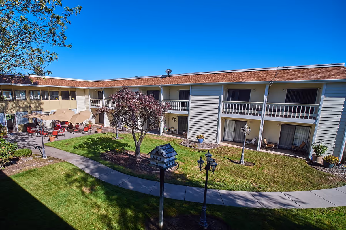 Outdoor courtyard area of a senior living facility with a two-story building surrounding a grassy lawn. The courtyard features a tree, birdhouse, lamp posts, and a seating area with tables, chairs, and umbrellas. The building has balconies and sliding glass doors leading to the courtyard.