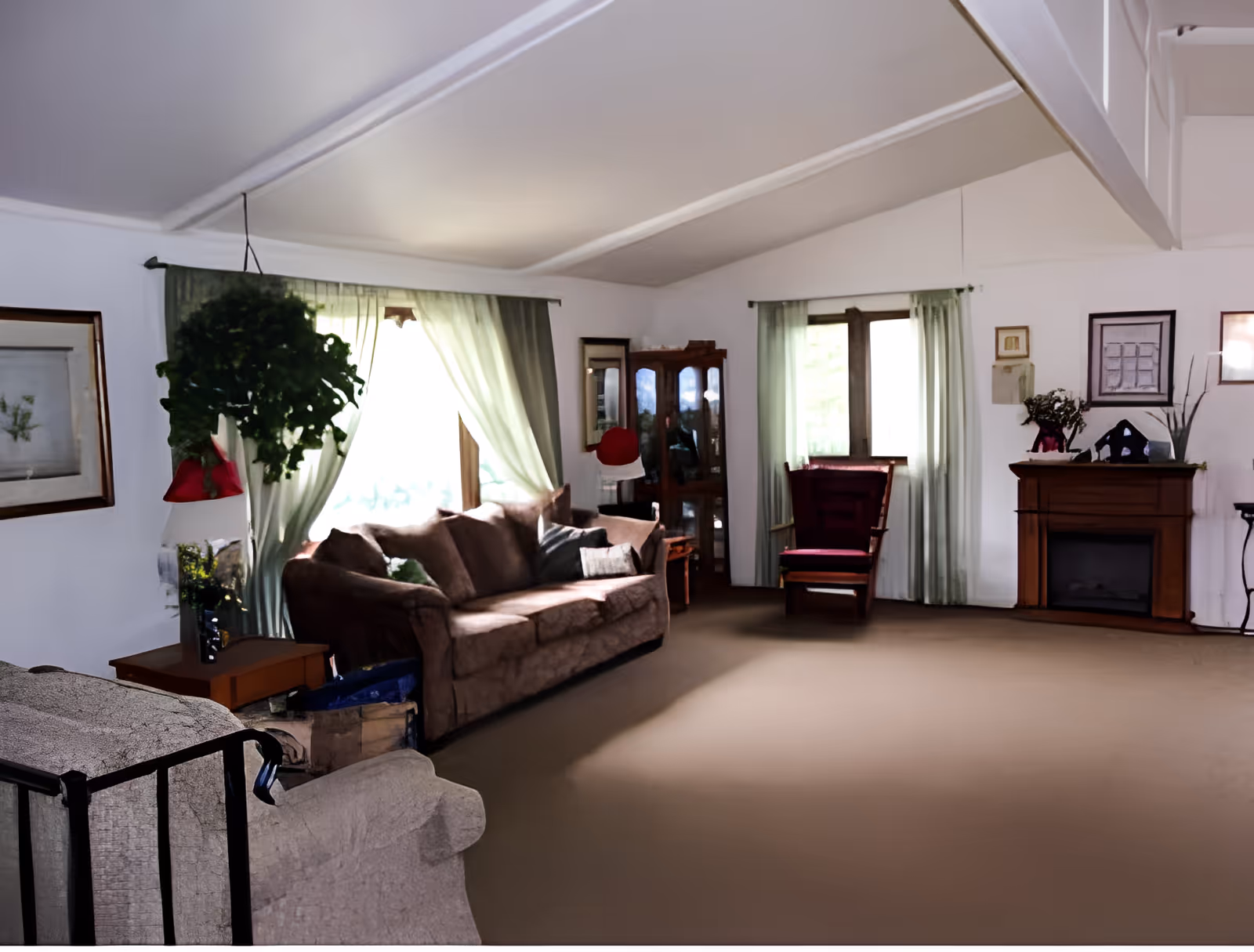 A spacious living room with beige carpet, a brown sofa, a red armchair, and a fireplace. The room has two windows with light green curtains, a hanging plant, framed pictures on the walls, and a wooden cabinet with glass doors.