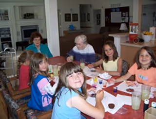 A group of six children sitting around a dining table in a well-lit room, smiling and engaging with each other. The table has plates, glasses, and some food items. The background shows a kitchen area and some wall decorations.