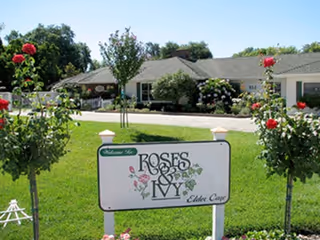 Exterior view of Roses & Ivy Elder Care facility with a green lawn, rose bushes, and a sign in the foreground displaying the facility's name. The building is a single-story structure with a gray roof and surrounded by trees and shrubs.