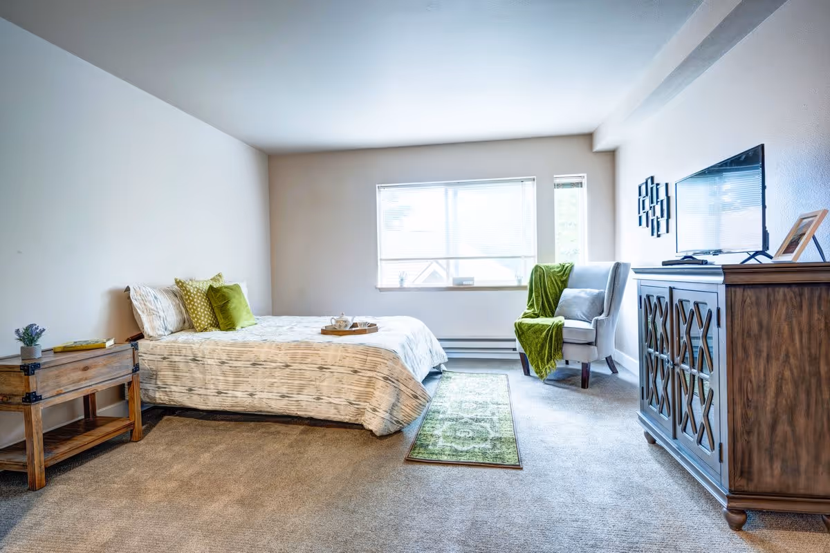 A bright bedroom with a bed against the left wall, decorated with green and beige pillows and a beige patterned bedspread. Next to the bed is a wooden nightstand with a small plant and a book. A green runner rug is placed on the carpeted floor leading to a window with blinds. On the right side, there is a gray armchair with a green throw blanket and a wooden dresser with a flat-screen TV and a framed photo on top. The walls are light-colored and the room is well-lit with natural light from the window.