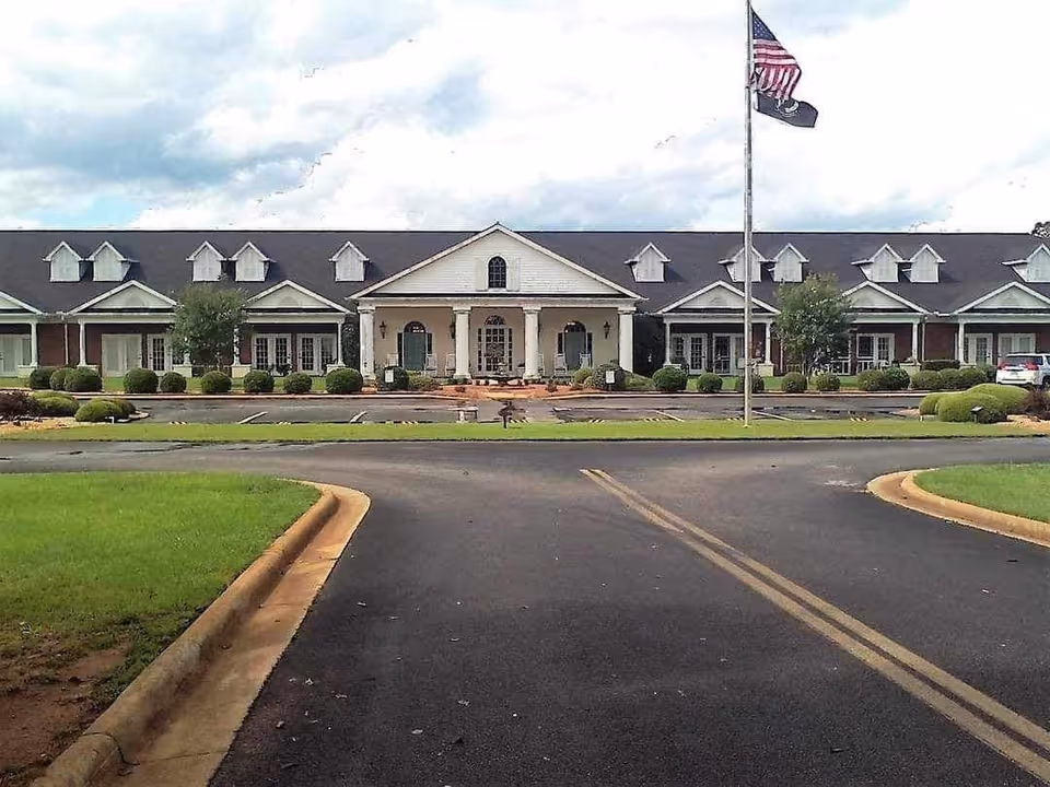 Front exterior view of Sumter Retirement Village building with a large driveway, manicured bushes, and an American flag on a flagpole in front.