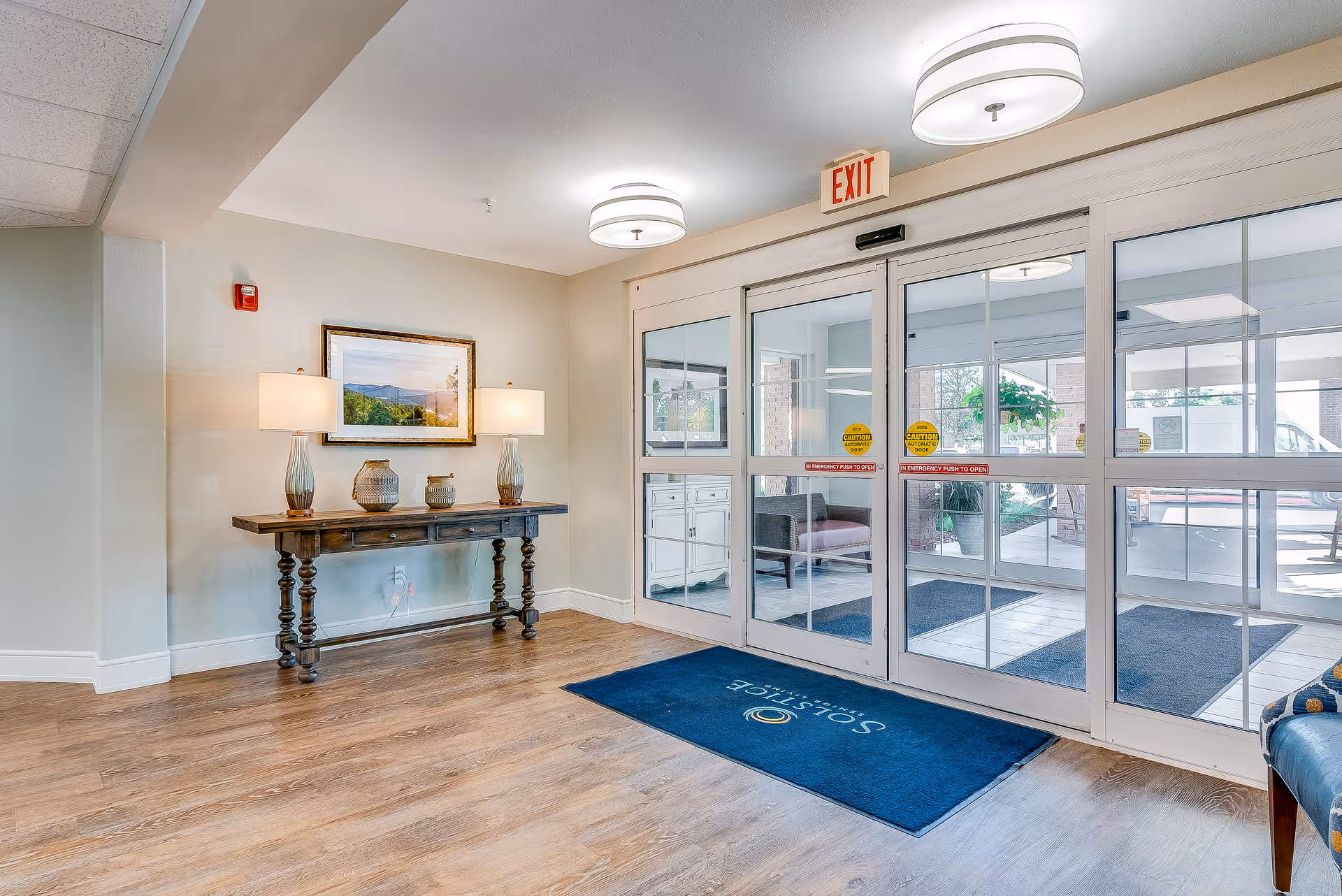 Entrance lobby with glass automatic doors, a console table with lamps and decorative vases, and a blue welcome mat.