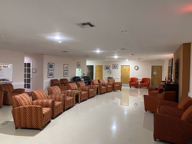 A spacious common area in an assisted living facility with multiple patterned armchairs arranged in rows along the walls. The room has a polished tile floor, white walls with framed pictures, and several doors leading to other rooms. The lighting is bright with ceiling lights, and there is a clock on one wall.