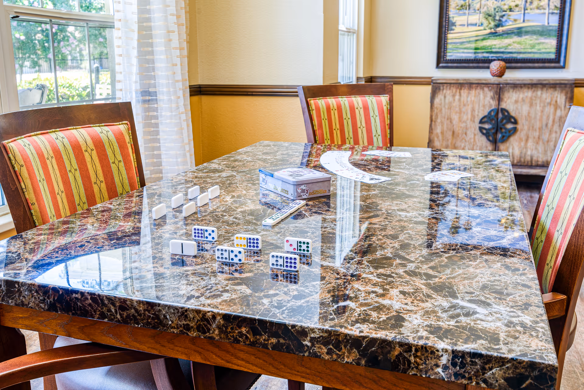 A polished marble table with a set of dominoes arranged on it, surrounded by wooden chairs with striped red and green cushions. The room has large windows with sheer curtains letting in natural light, and a wooden cabinet with a framed picture above it is visible in the background.