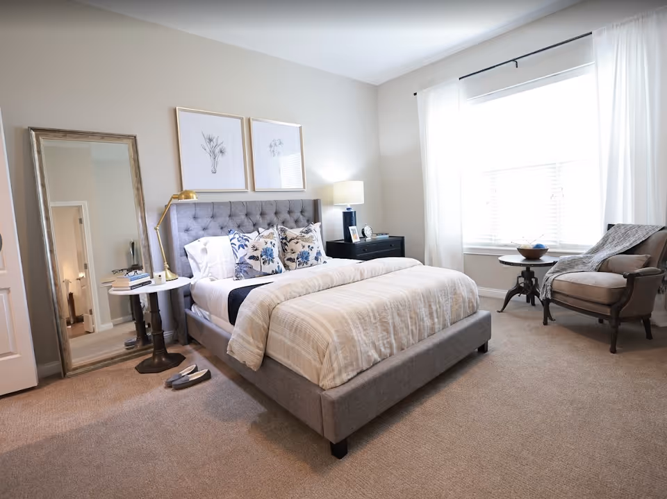 Well-lit bedroom with a gray upholstered bed, bedside tables, a floor mirror, and an armchair by a large window.