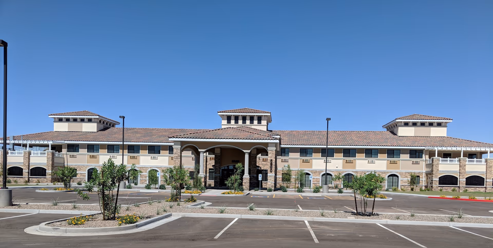 Front exterior view of The Mission at Agua Fria Senior Living building with a large covered entrance, multiple windows, and a tiled roof under a clear blue sky. The parking lot and landscaped areas with small trees and plants are visible in the foreground.