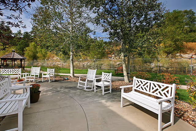 Outdoor patio with white benches and rocking chairs, trees, a gazebo, and a fenced landscaped yard.