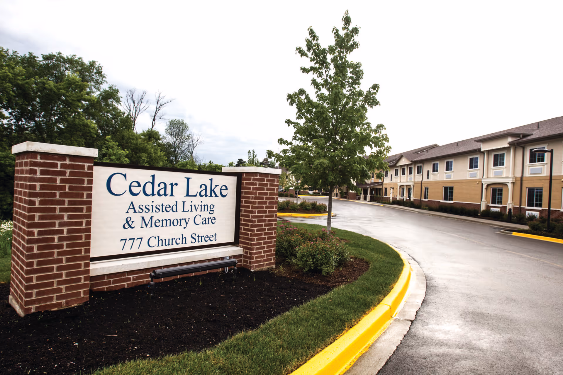 Entrance sign for Cedar Lake Assisted Living & Memory Care at 777 Church Street with a two-story building and driveway in the background, surrounded by green trees and landscaping.