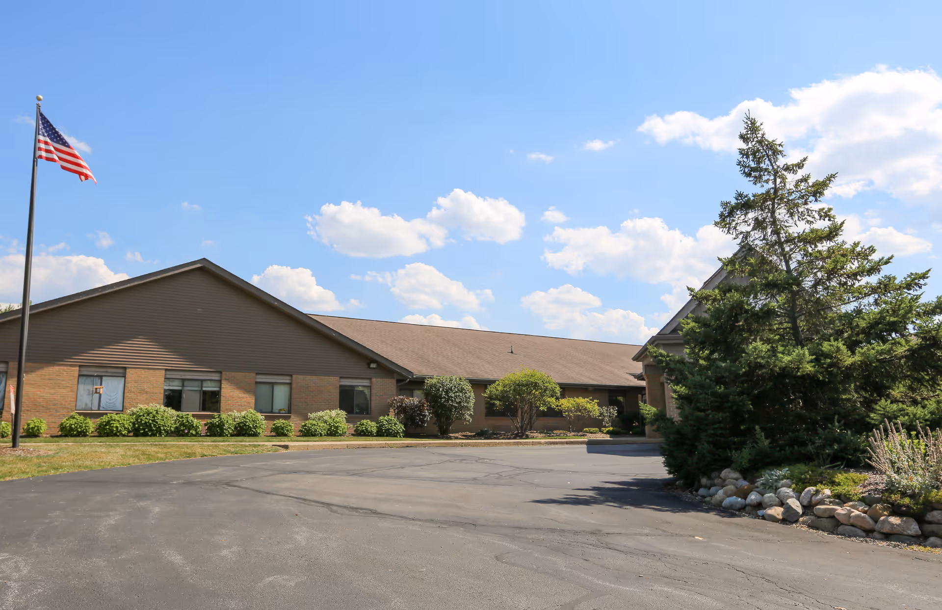 Exterior view of a single-story brick building with a sloped roof, surrounded by shrubs and trees. An American flag is flying on a flagpole to the left, and the sky is partly cloudy with blue patches.