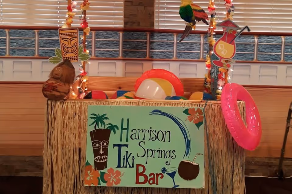 Tiki-themed indoor bar setup with a sign reading 'Harrison Springs Tiki Bar', beach balls, a pink inflatable ring, and tropical decorations.