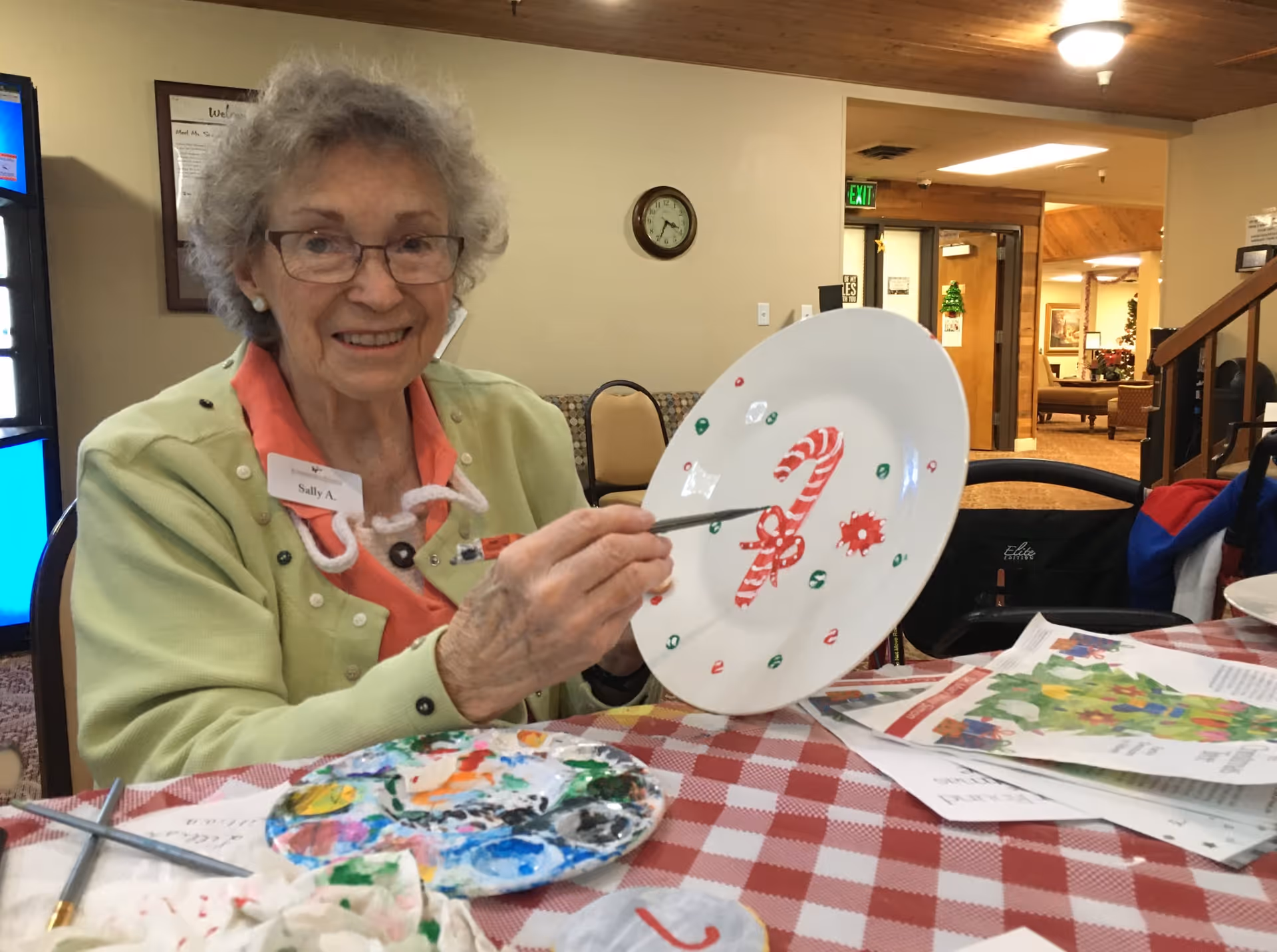 An elderly woman with gray hair and glasses is sitting at a table covered with a red and white checkered tablecloth. She is smiling and holding a white plate decorated with a painted candy cane and other festive designs. There are paint supplies and papers on the table. The background shows a cozy interior with chairs, a clock on the wall, and an open doorway leading to another room.