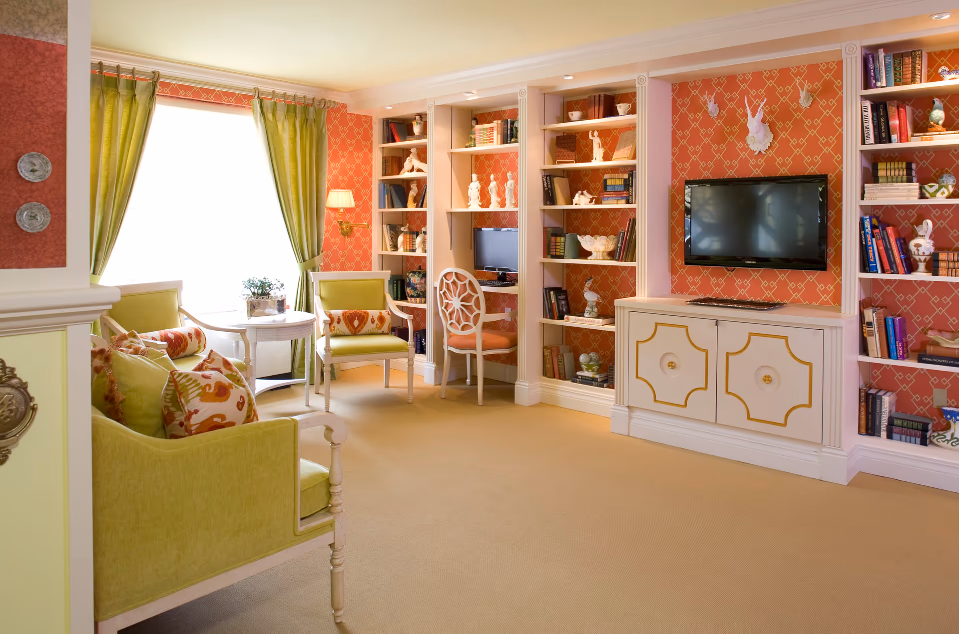 Bright cozy living room with green upholstered seating, built-in bookshelves, a wall-mounted TV, and patterned coral wallpaper.