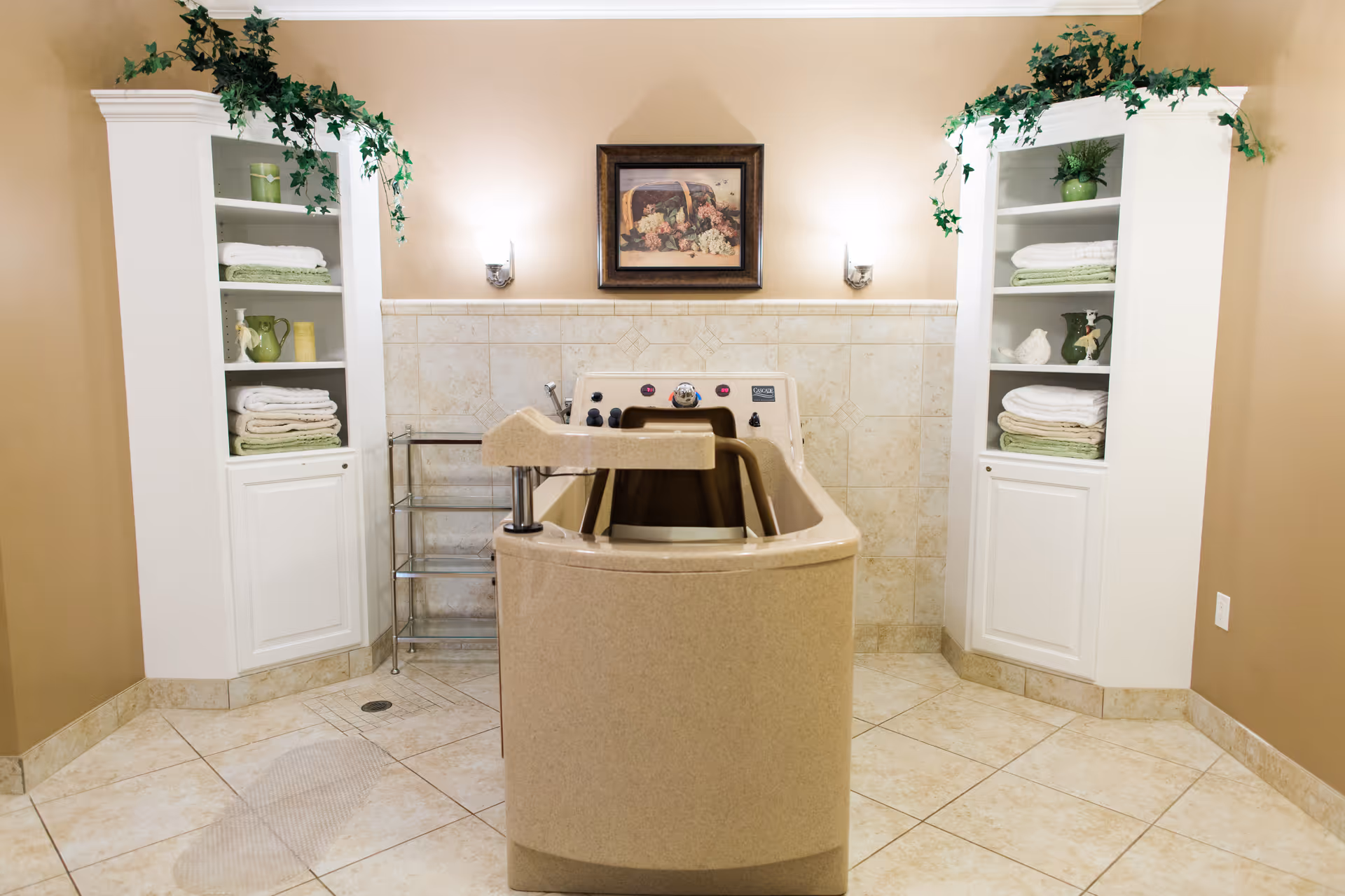 A beige-colored walk-in bathtub in a tiled bathroom with two white shelving units on either side, each holding folded towels and decorative items. There are green plants on top of the shelves, wall-mounted lights, and a framed picture above the bathtub.