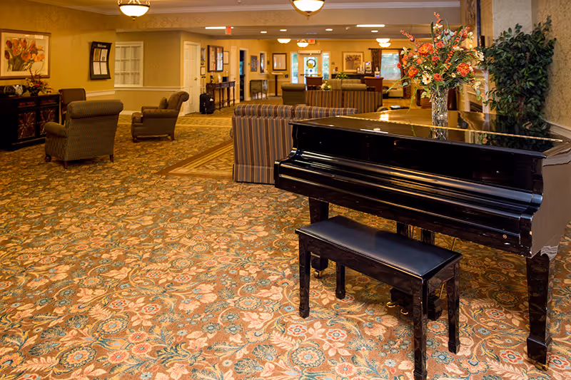 A spacious senior living facility common area with a patterned carpet, a black grand piano with a matching bench, a vase of flowers on the piano, several upholstered chairs and sofas, warm lighting, and decorative plants and artwork on the walls.