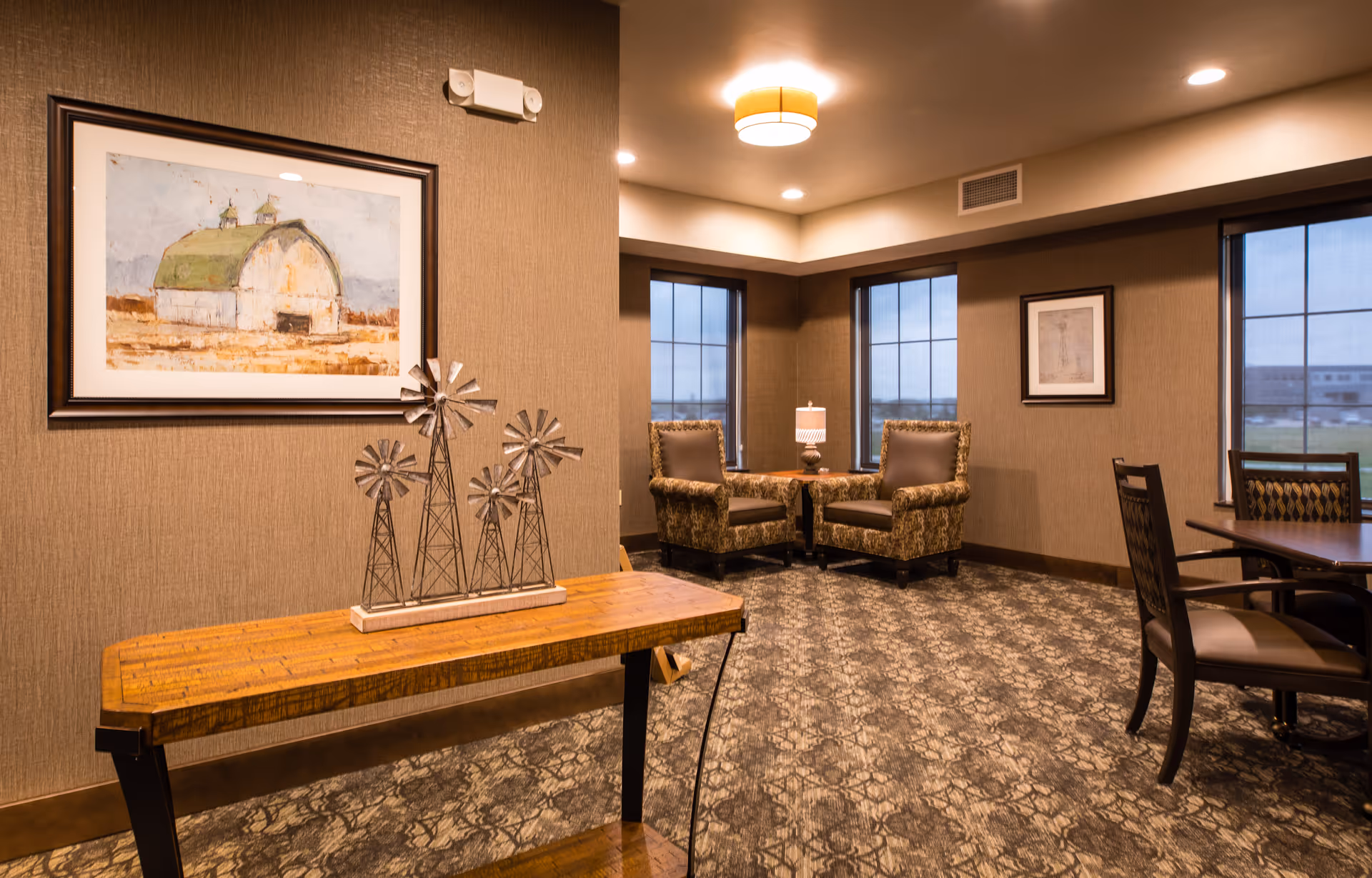 Stylish senior living common room with upholstered armchairs by windows, a wooden console table topped with metal windmill sculptures, framed artwork, and a dining table.