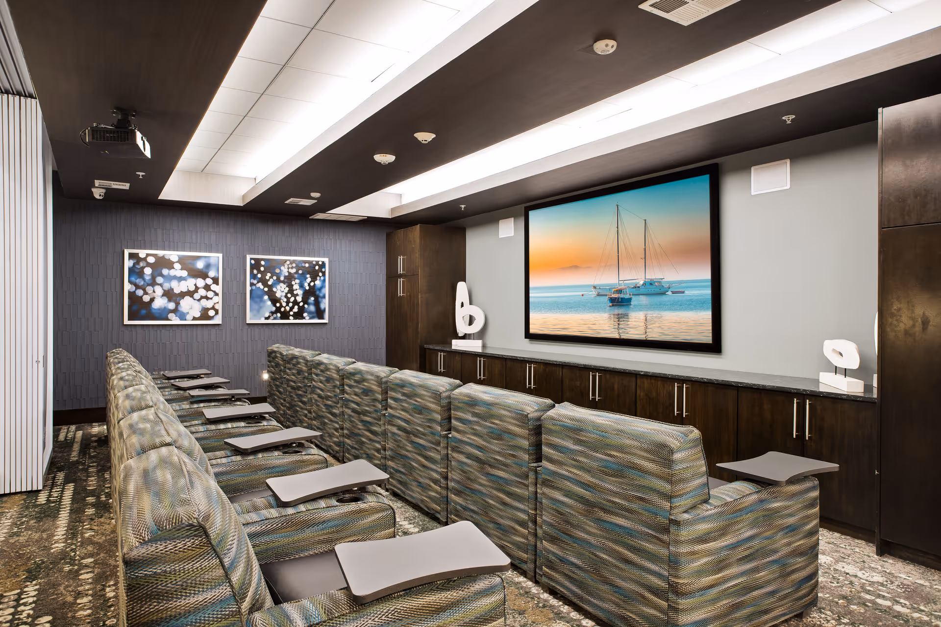 A small theater room with two rows of patterned reclining chairs, each equipped with a small attached table. At the front, there is a large screen displaying an image of sailboats on calm water during sunset. The room has dark wood cabinetry beneath the screen, modern ceiling lights, and two abstract sculptures on the cabinet. Two framed abstract photos with bokeh lights hang on the side wall.
