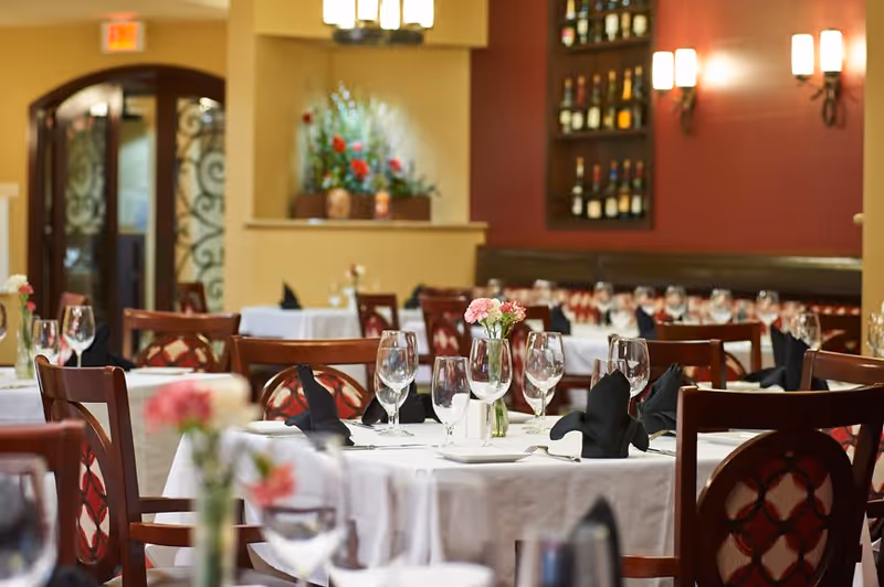 Elegant dining room with round tables covered in white tablecloths, each set with wine glasses, black folded napkins, and small flower vases. The room features wooden chairs with patterned backs, warm lighting, and a wall with shelves displaying bottles of wine.