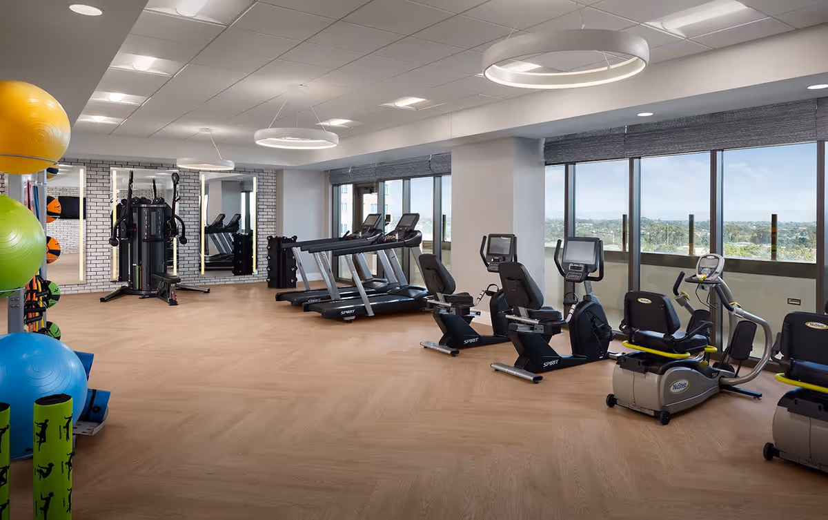 A bright and spacious fitness room with large windows offering a scenic view. The room contains various exercise equipment including treadmills, recumbent bikes, and a multi-station gym machine. Colorful exercise balls and foam rollers are visible on the left side. The floor has a light wood finish and modern circular ceiling lights illuminate the space.