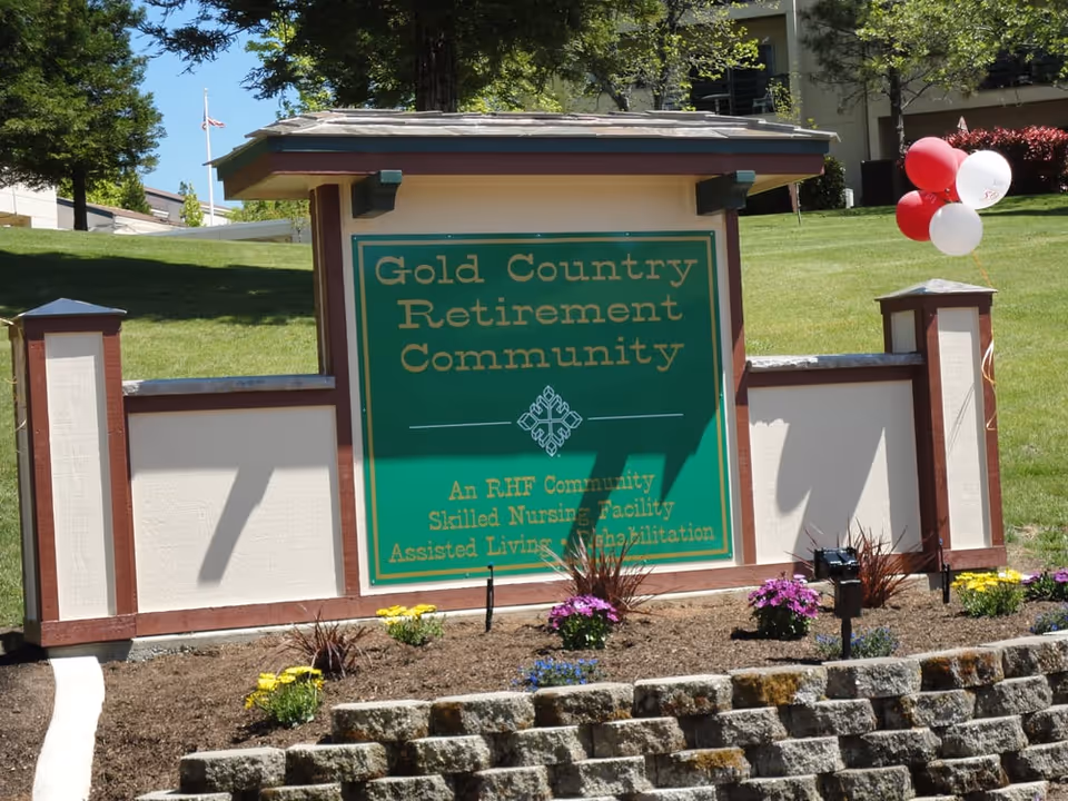 A landscaped entrance sign reading "Gold Country Retirement Community" with balloons and flowers on a grassy lawn.
