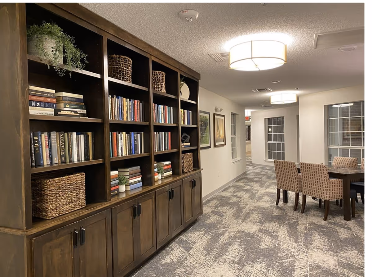 Interior view of a senior living facility hallway with a large wooden bookshelf filled with books, baskets, and decorative plants on the left. On the right, there is a dining table with four patterned chairs. The space is well-lit with ceiling lights and has carpeted flooring with a modern pattern.