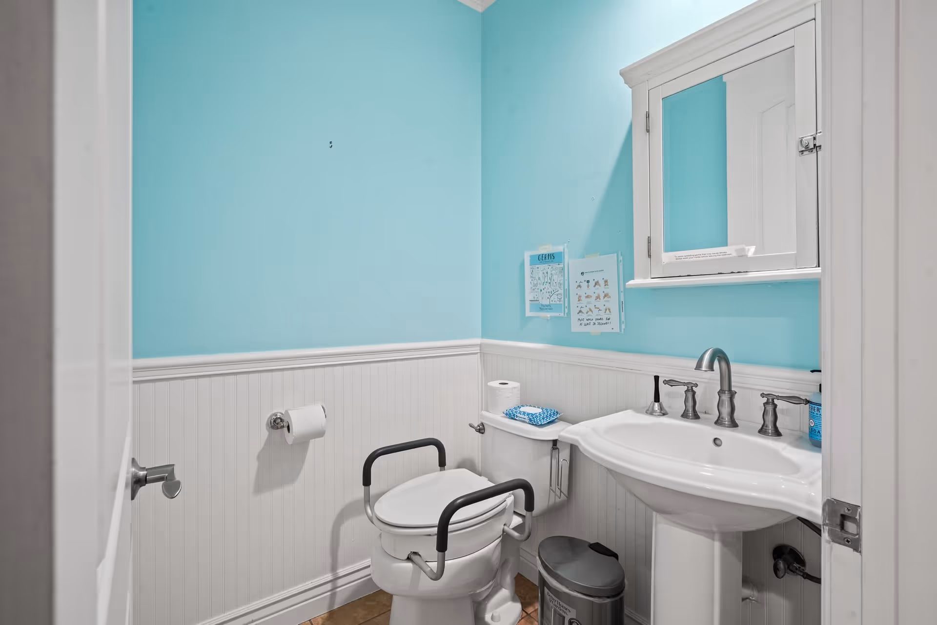A small bathroom with light blue walls and white wainscoting. It features a white toilet with black safety rails, a white pedestal sink with silver faucets, a mirrored medicine cabinet above the sink, and a small trash can on the floor. There are two informational posters on the wall above the toilet, and a roll of toilet paper is mounted on the wall.
