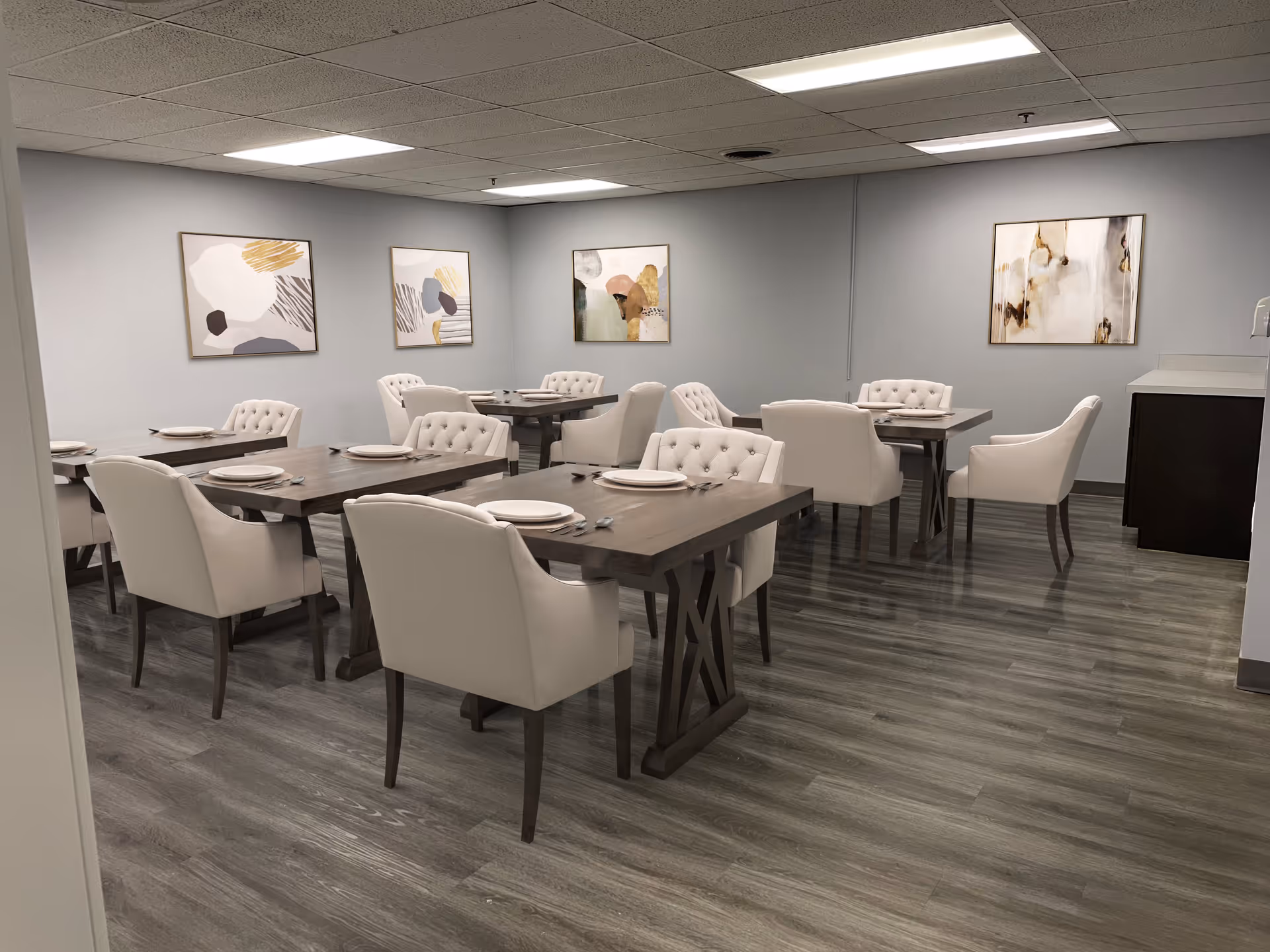 A dining room with several wooden tables set with white plates and silverware. Each table is surrounded by white cushioned chairs. The room has light gray walls adorned with abstract art paintings and a wood-patterned floor. The ceiling has recessed lighting panels.