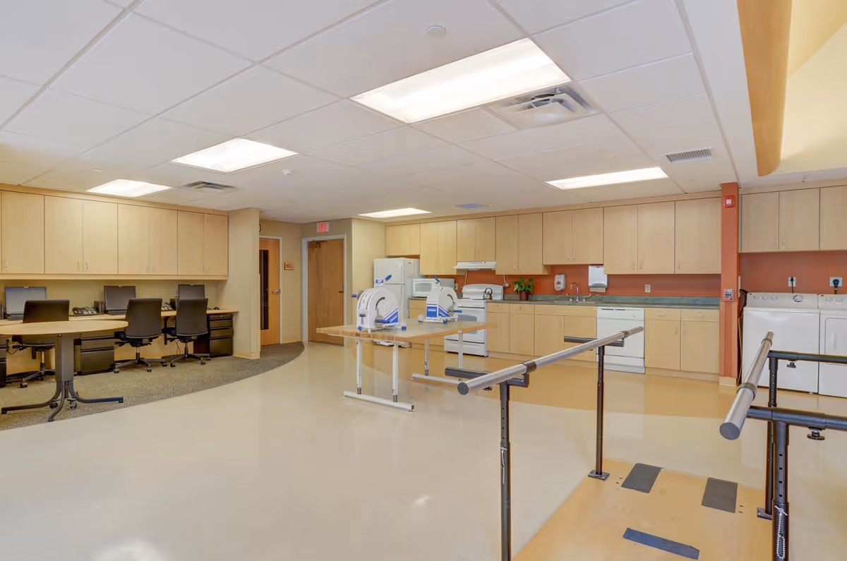 A spacious room with a kitchen area featuring light wood cabinets, a refrigerator, stove, dishwasher, and washing machines. There are two tables with medical or therapy equipment on them and parallel bars for walking exercises in the foreground. On the left side, there is a workstation with three office chairs and computers. The room has a clean, well-lit appearance with a tiled floor and drop ceiling lights.