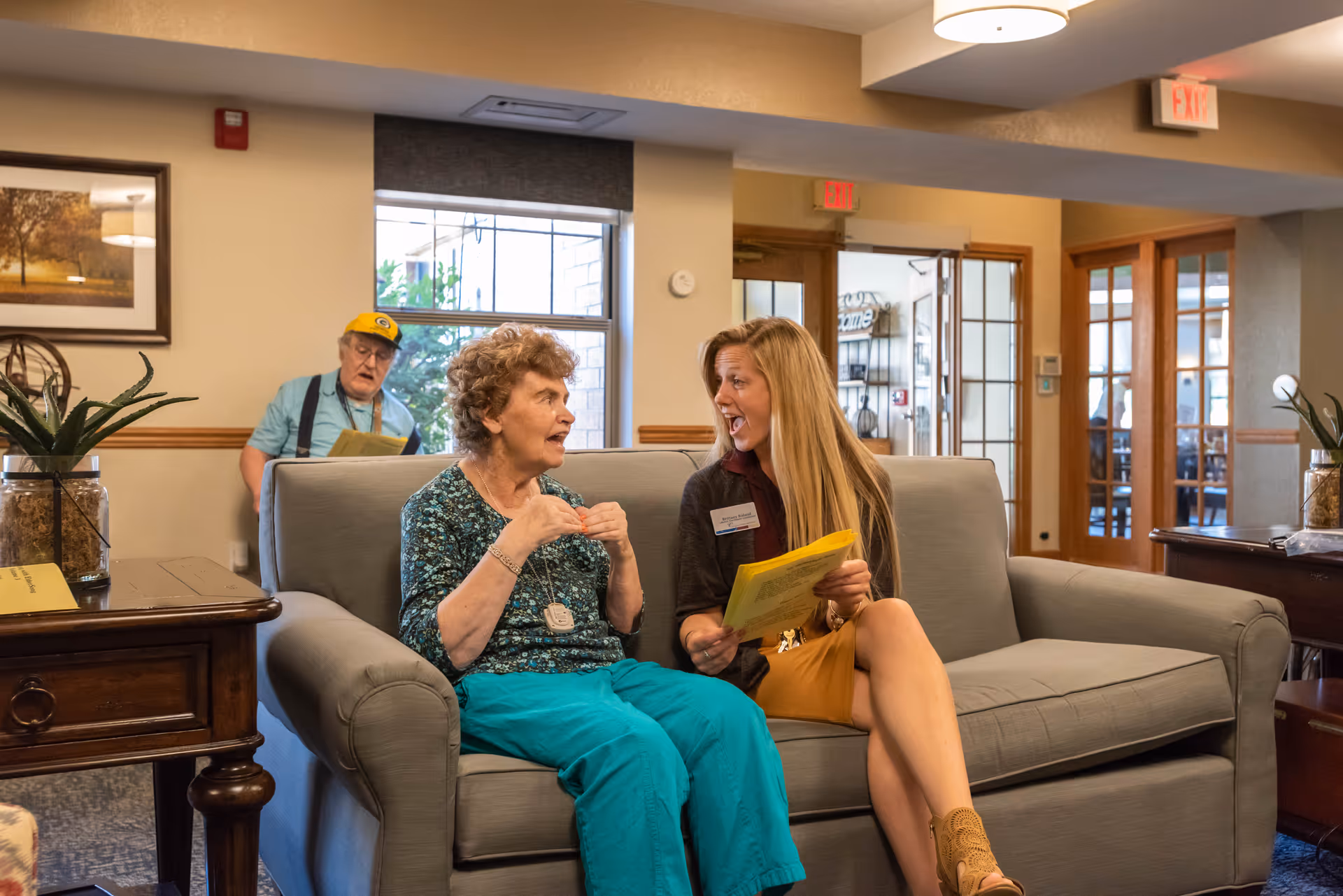 Two women sit and talk on a couch in an assisted living facility's common lounge, with a man seated in the background.