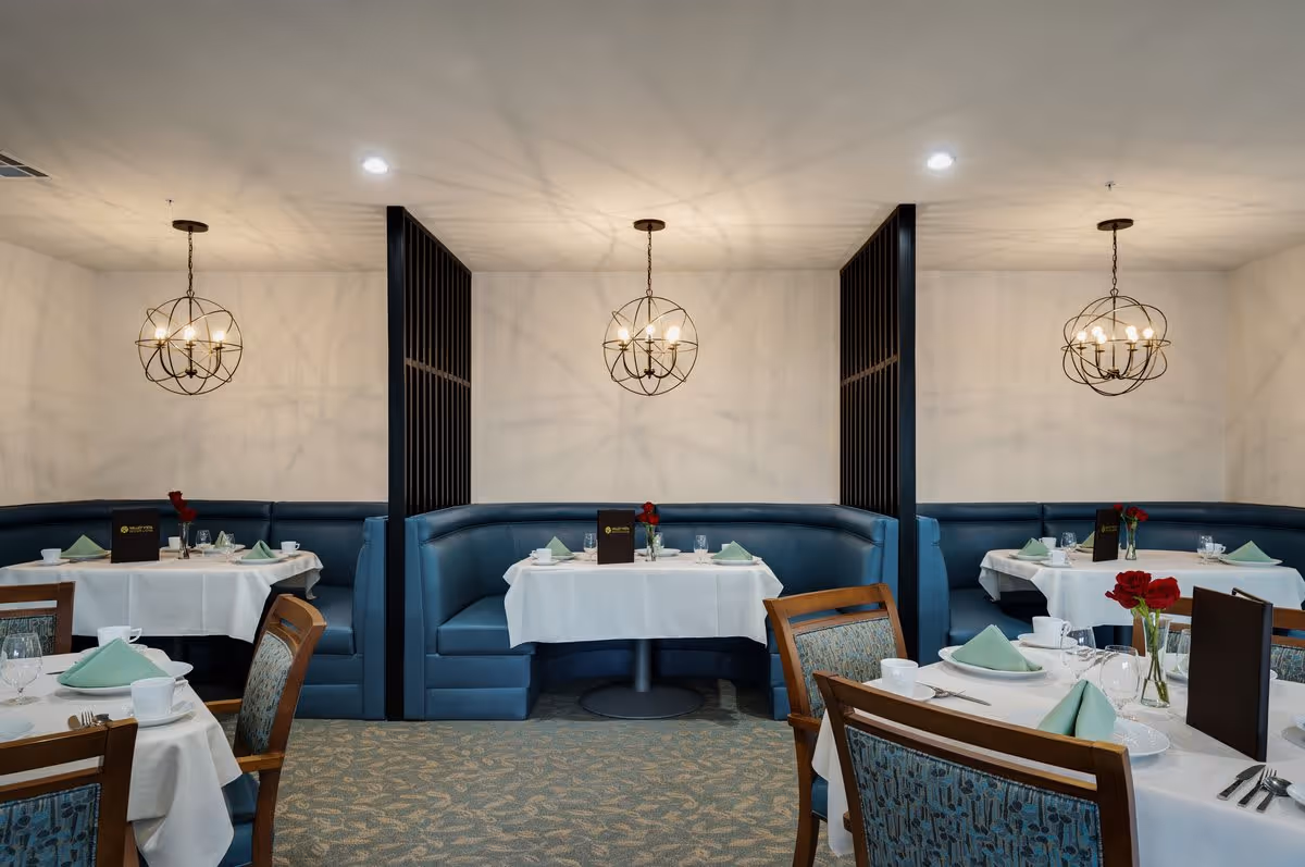 Dining area with three semi-circular blue booths separated by black partitions, each booth has a table set with white tablecloths, green folded napkins, white cups, glasses, and a menu. There are also wooden chairs with patterned upholstery around other tables. Three spherical chandeliers hang from the ceiling, casting shadows on the walls.
