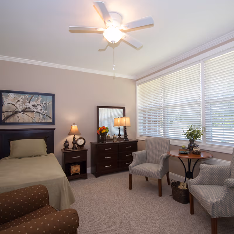 A cozy bedroom with a single bed covered in a green bedspread, a nightstand with a lamp and clock, a dresser with a mirror and two lamps, two upholstered armchairs, a round wooden table with decorative items, and large windows with white blinds letting in natural light.