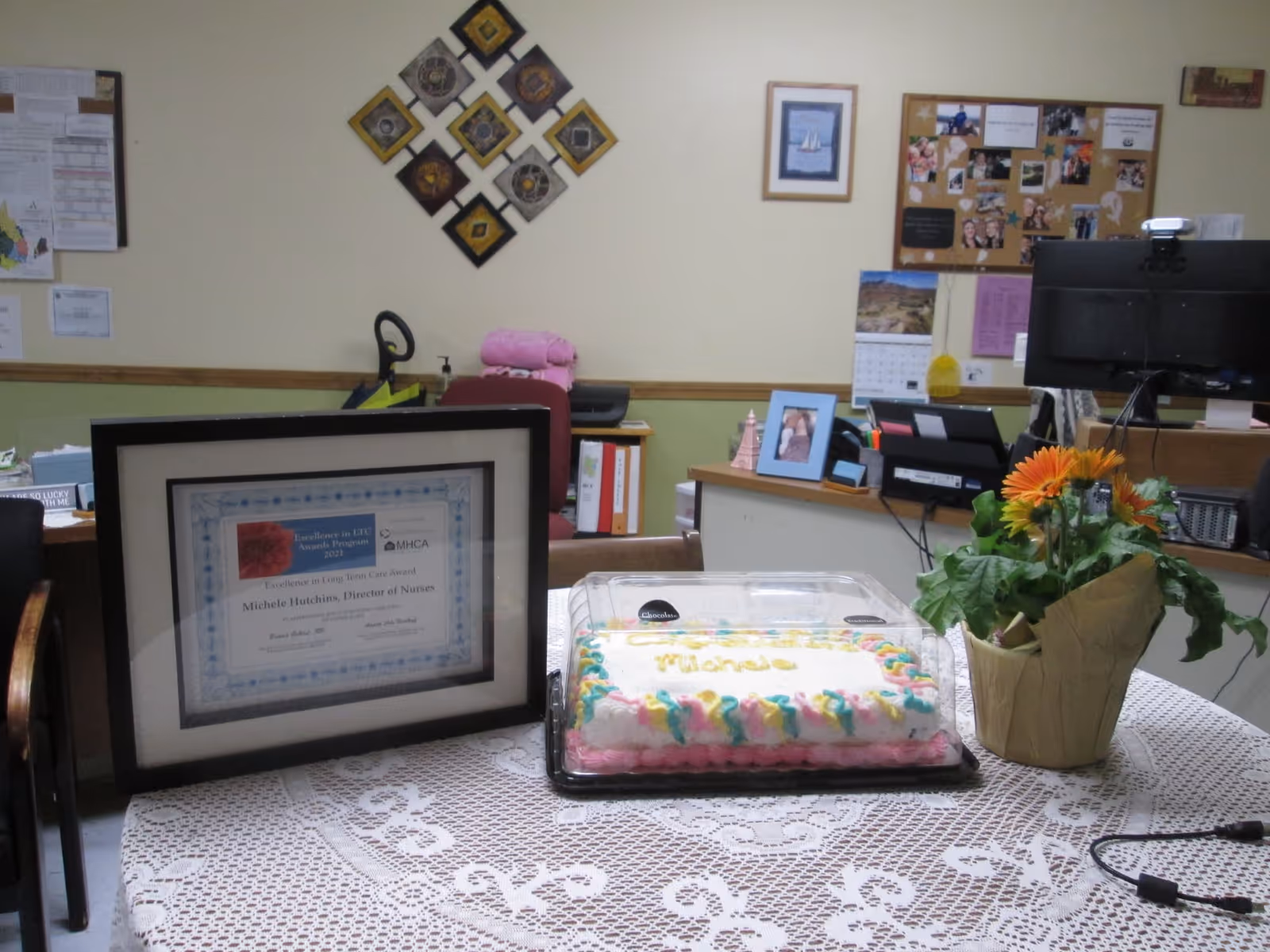 A reception/office interior with a lace-covered table holding a framed certificate, a plastic-covered sheet cake and a potted flower, with desks and a bulletin board in the background.