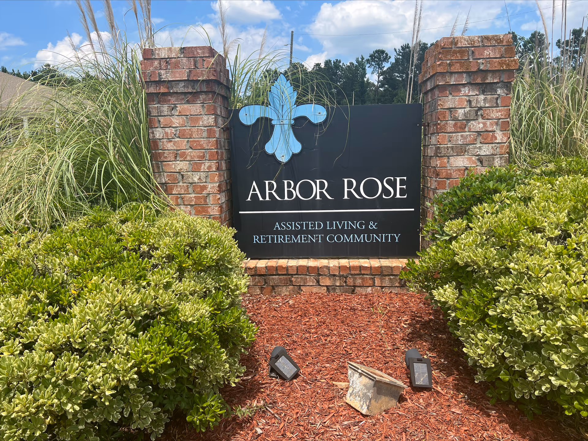 Outdoor sign for Arbor Rose Assisted Living & Retirement Community surrounded by green bushes and tall grass with a blue sky and trees in the background.