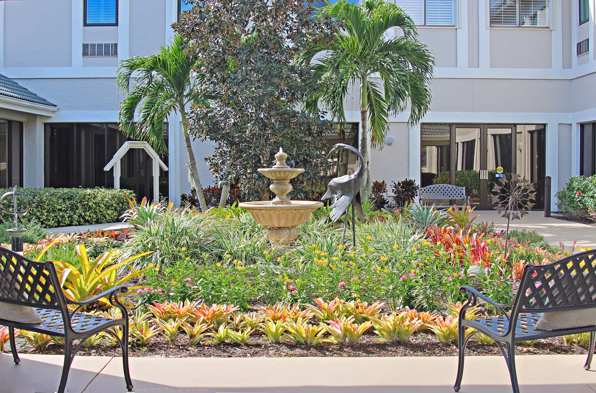 A landscaped outdoor courtyard at Arbor Trace featuring a central two-tiered stone fountain surrounded by lush greenery, colorful flowers, palm trees, and a metal bird sculpture. There are two black metal benches with cushions facing the garden area, and the building with large windows and doors is visible in the background.