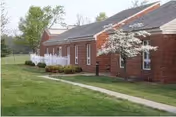 Exterior view of a single-story brick building with a sloped roof, white-framed windows, and a small white flowering tree in front. There is a concrete walkway leading alongside the building and a grassy lawn surrounding the area.