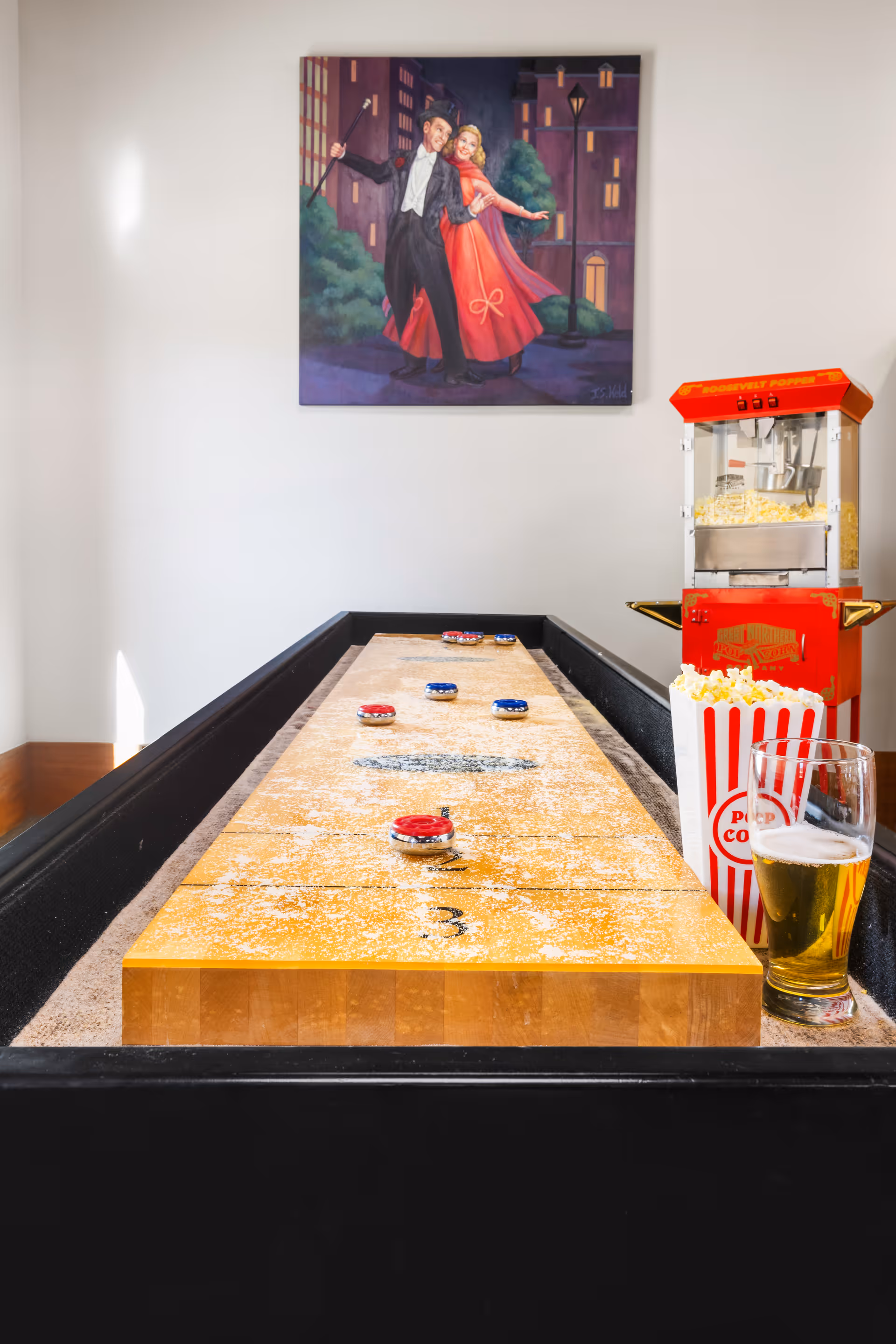 A shuffleboard table with red and blue pucks, a glass of beer, and a striped popcorn container filled with popcorn. A red popcorn machine is visible in the background, and a painting of a dancing couple in formal attire hangs on the wall above.