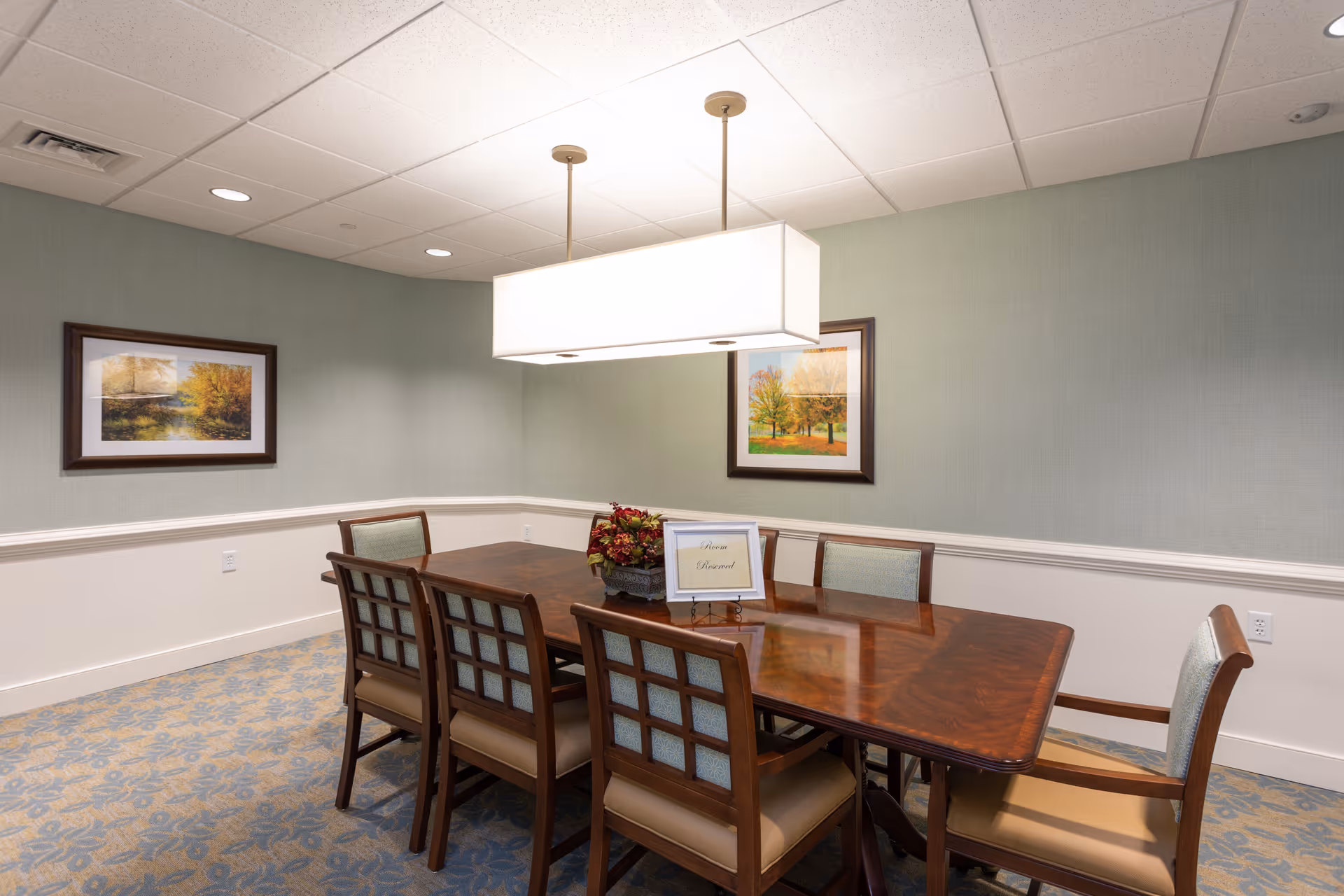 A reserved dining room with a rectangular wooden table surrounded by eight cushioned chairs. The room has light green walls with white wainscoting, two framed landscape paintings, a floral centerpiece on the table, and a large rectangular ceiling light fixture above the table.