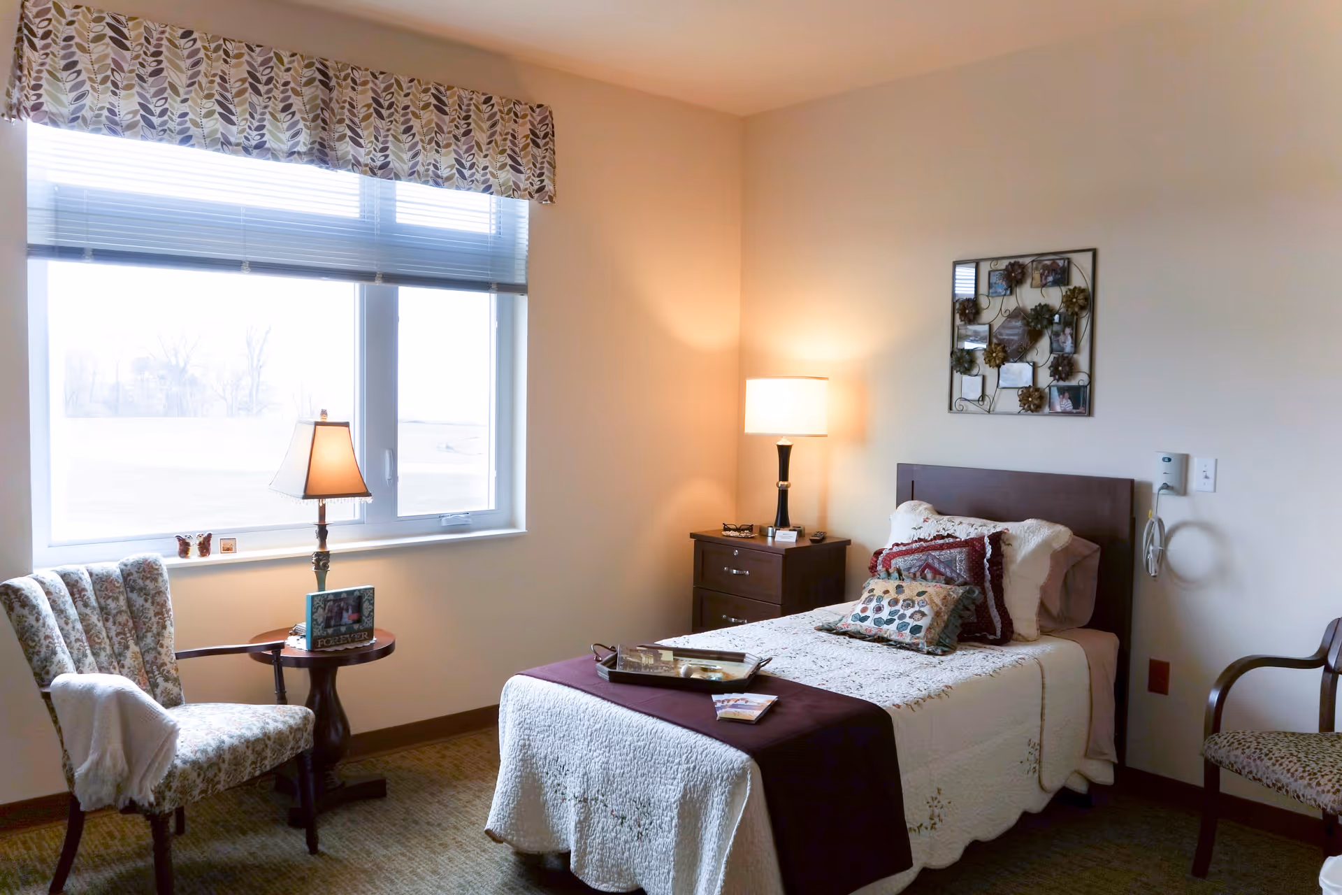 A cozy bedroom in a senior living facility with a single bed covered in a white quilt and decorative pillows. There is a wooden nightstand with a lamp and some items on it next to the bed. A window with blinds and a patterned valance lets in natural light. Two upholstered chairs with floral patterns and a small round table with a lamp and framed photo are near the window. A decorative wall hanging with photos is above the bed.