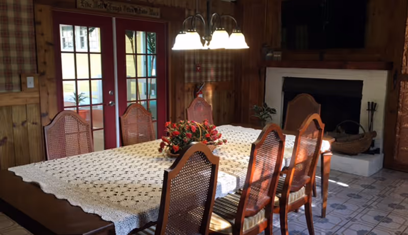 A dining room with a wooden table covered by a white lace tablecloth and surrounded by six wooden chairs with woven backs. A floral centerpiece is placed on the table. The room features wood-paneled walls, a fireplace with a basket of firewood, and a set of double glass doors with red frames leading outside. A chandelier with five lights hangs above the table.