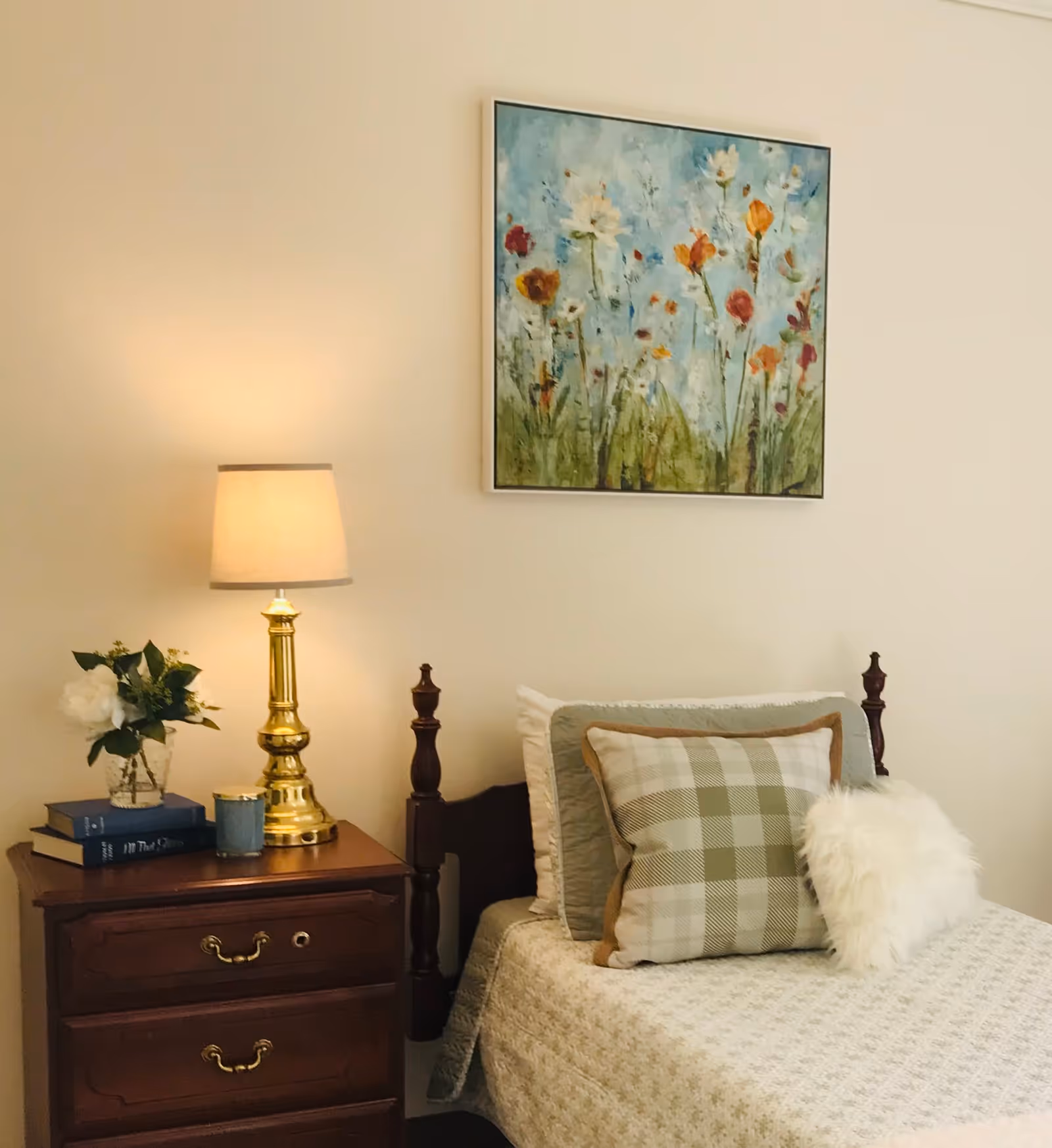 A cozy bedroom corner featuring a wooden bed with a beige quilt, a plaid pillow, and a fluffy white pillow. Next to the bed is a wooden nightstand with two drawers, holding a brass lamp with a beige lampshade, a small vase with white flowers, two books, and a blue candle. Above the bed hangs a colorful painting of flowers against a blue sky.