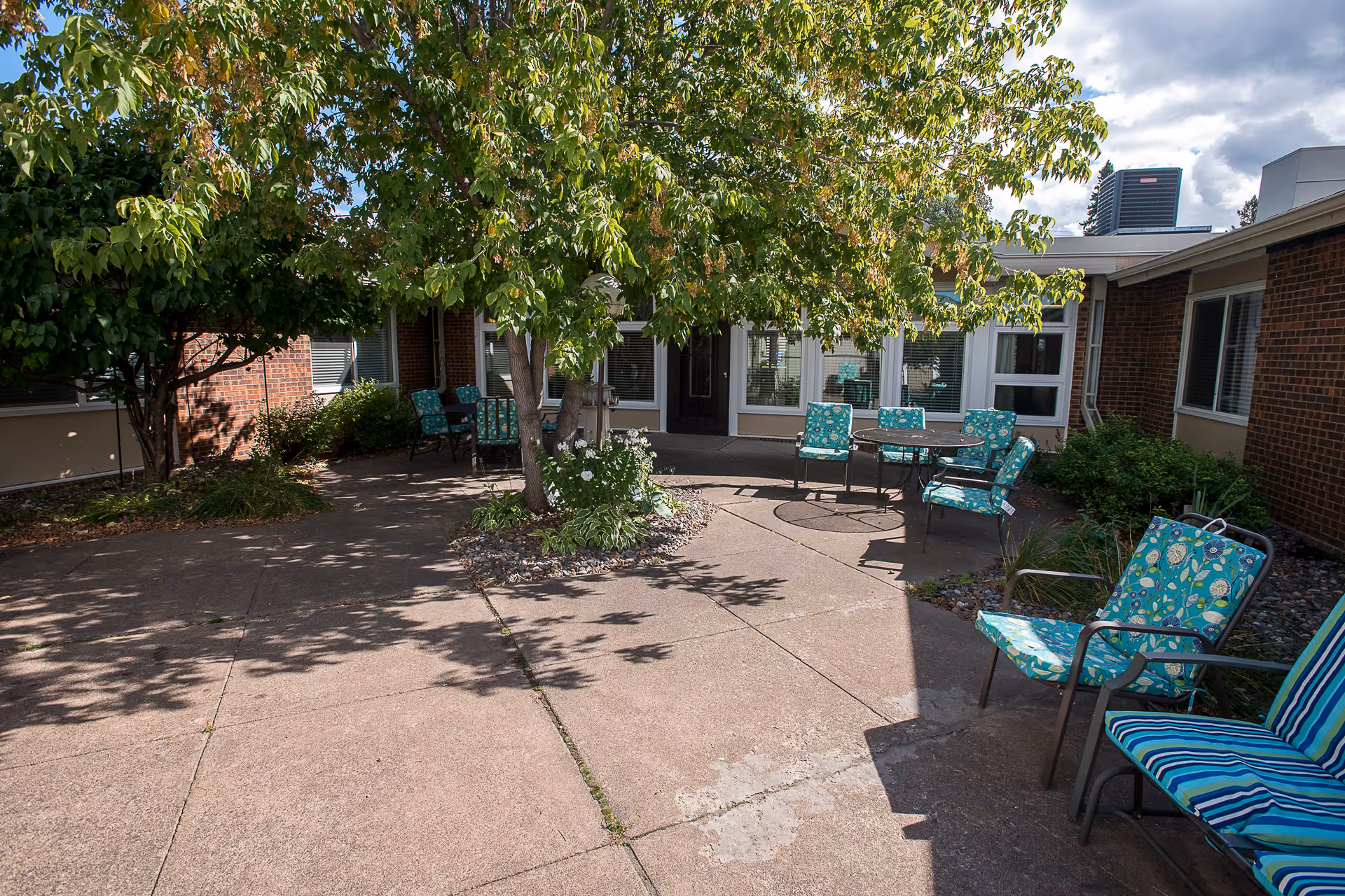 Sunlit courtyard with patio chairs and tables under a tree outside a single-story brick senior living building.