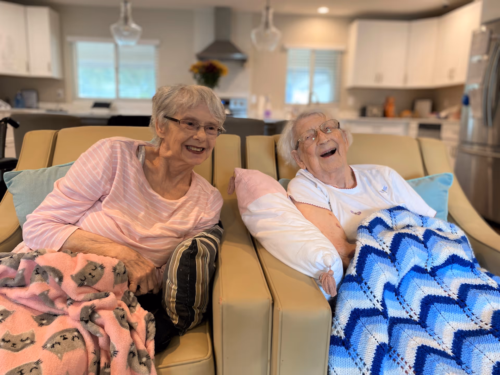 Two elderly women sitting on a beige couch in a cozy living room. Both are smiling and covered with colorful blankets, one pink with cat patterns and the other blue and white. The background shows a modern kitchen with white cabinets and stainless steel appliances.
