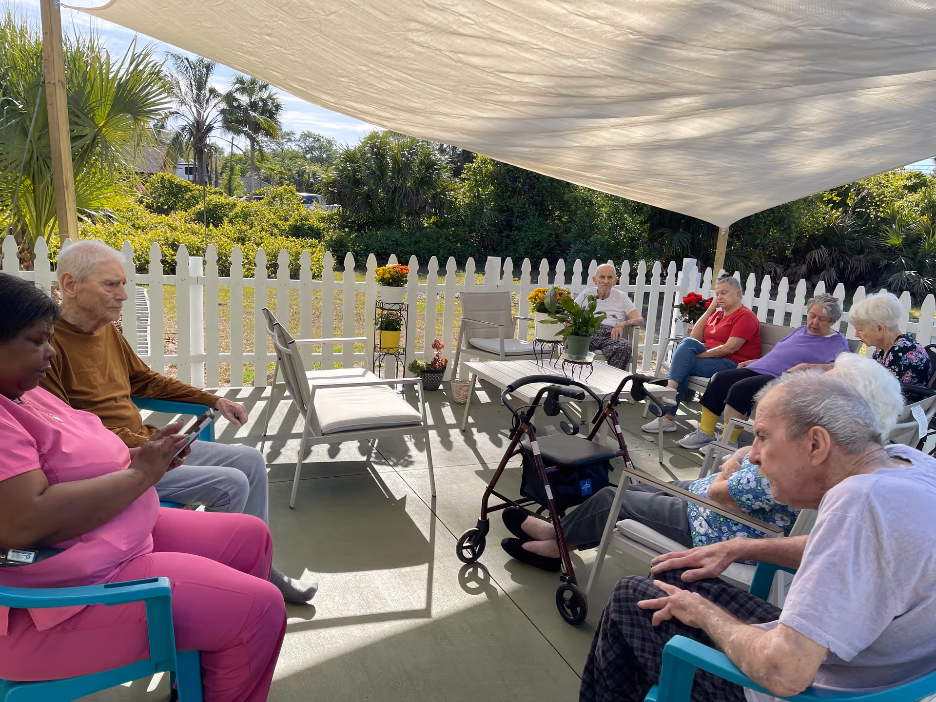 A group of elderly people and a caregiver sitting outdoors under a large white canopy. They are seated in a semi-circle on chairs around a small table with potted plants. The area is enclosed by a white picket fence with greenery and palm trees visible in the background.