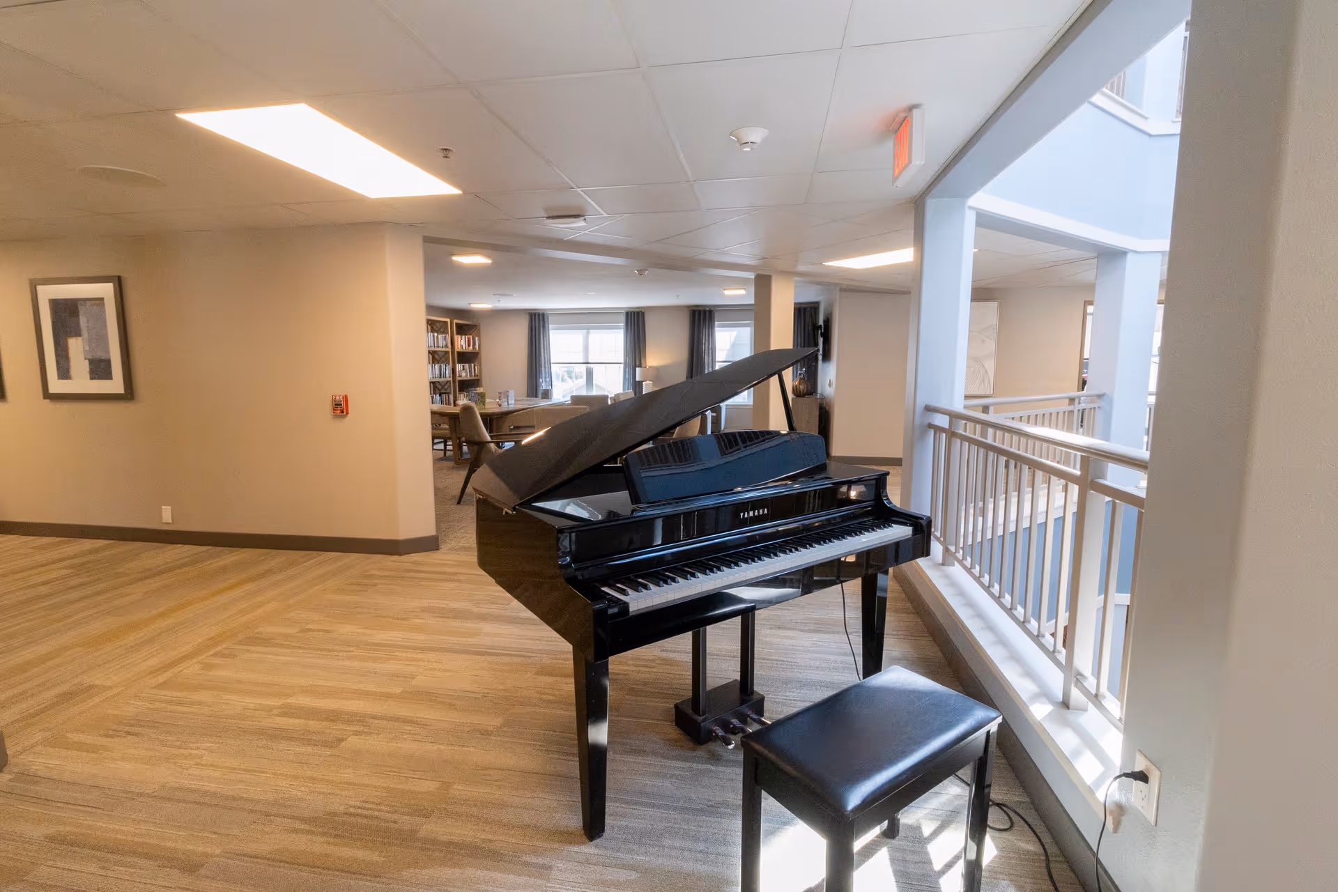 Interior view of a senior living facility common area featuring a black grand piano with an open lid and a matching bench. The space has light wood flooring, beige walls, and a railing overlooking a lower level. In the background, there are chairs, tables, bookshelves, and windows with curtains allowing natural light to enter.