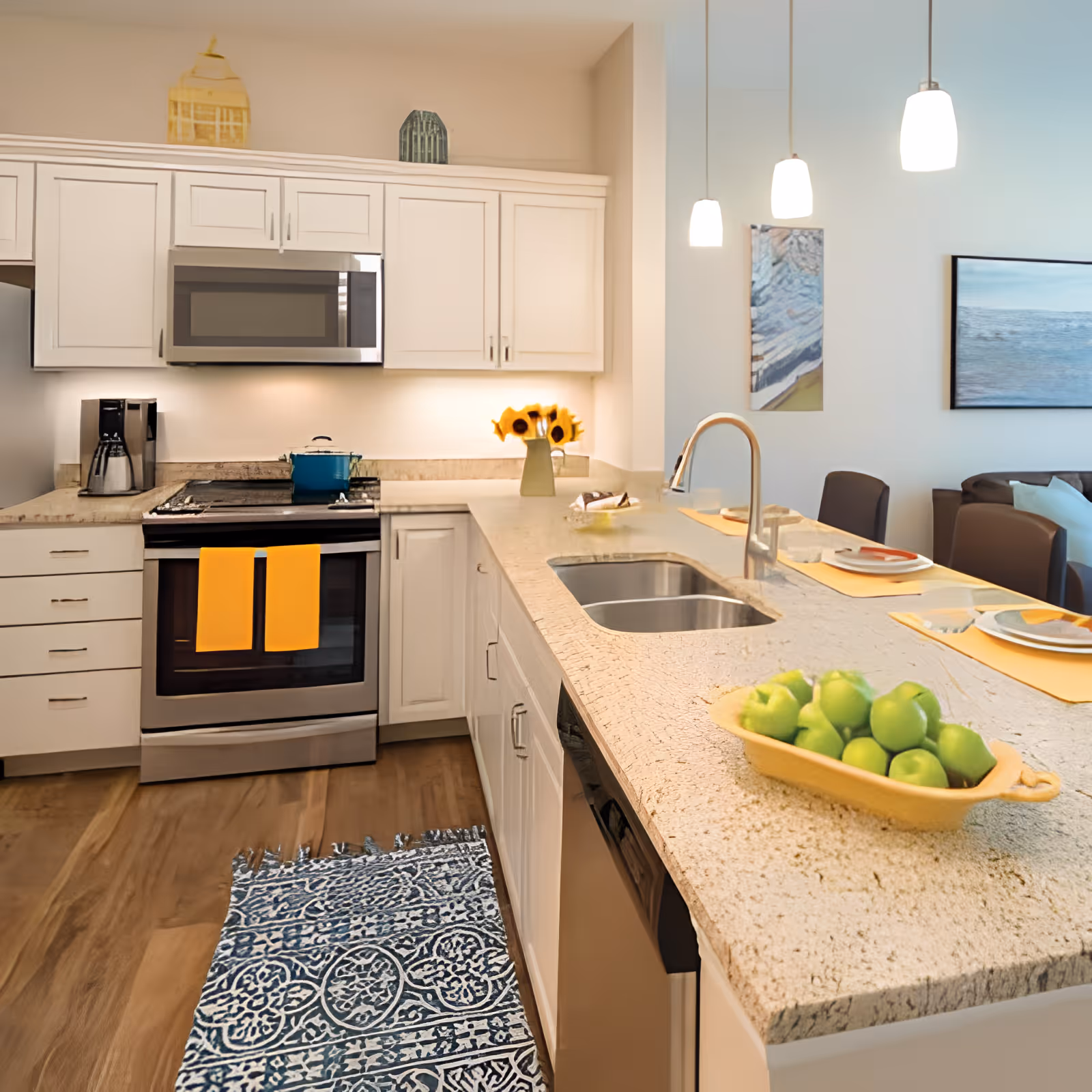 Bright modern kitchen with white cabinets, a granite island with sink and a bowl of green apples, stainless appliances and pendant lights.