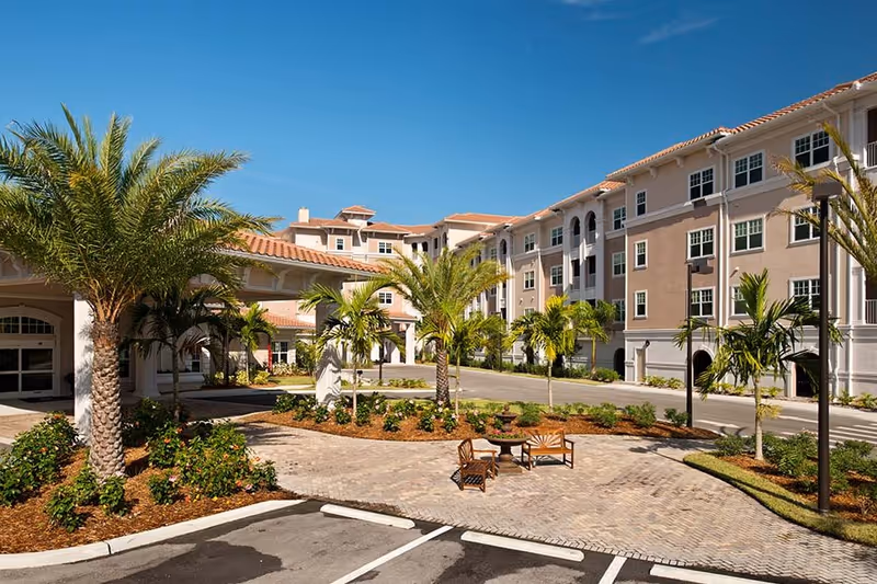 Exterior view of Diamond Oaks Village senior living facility featuring a multi-story building with beige and white walls, red-tiled roofs, palm trees, landscaped garden beds, a paved courtyard with outdoor seating, and a clear blue sky.