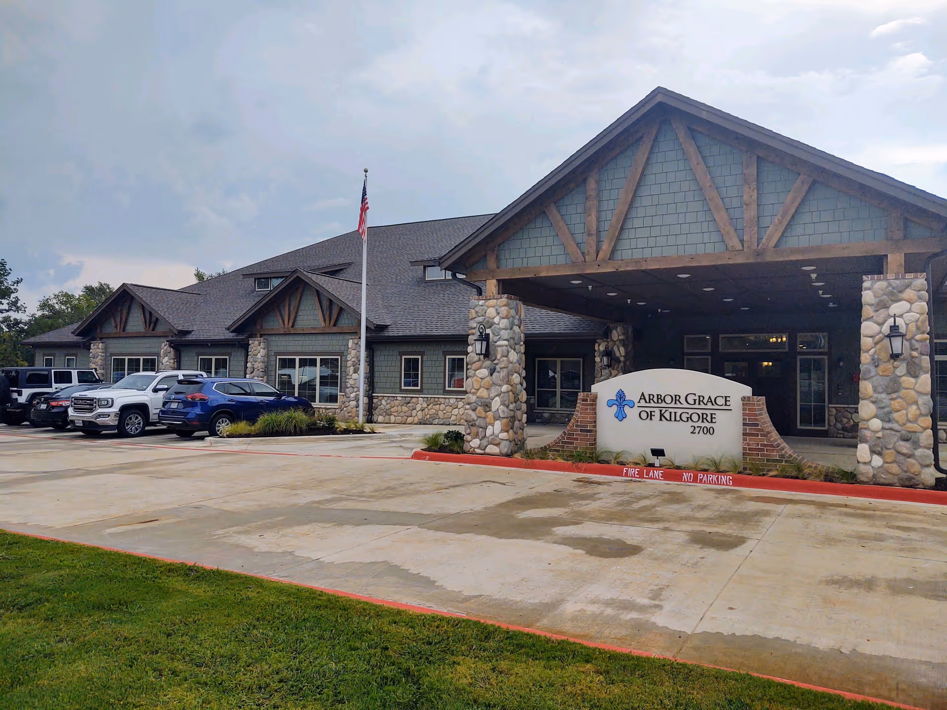 Exterior view of Arbor Grace of Kilgore senior living facility showing a large building with stone and wood accents, a covered entrance supported by stone pillars, an American flag on a flagpole, several parked cars, and a sign with the facility name and address.