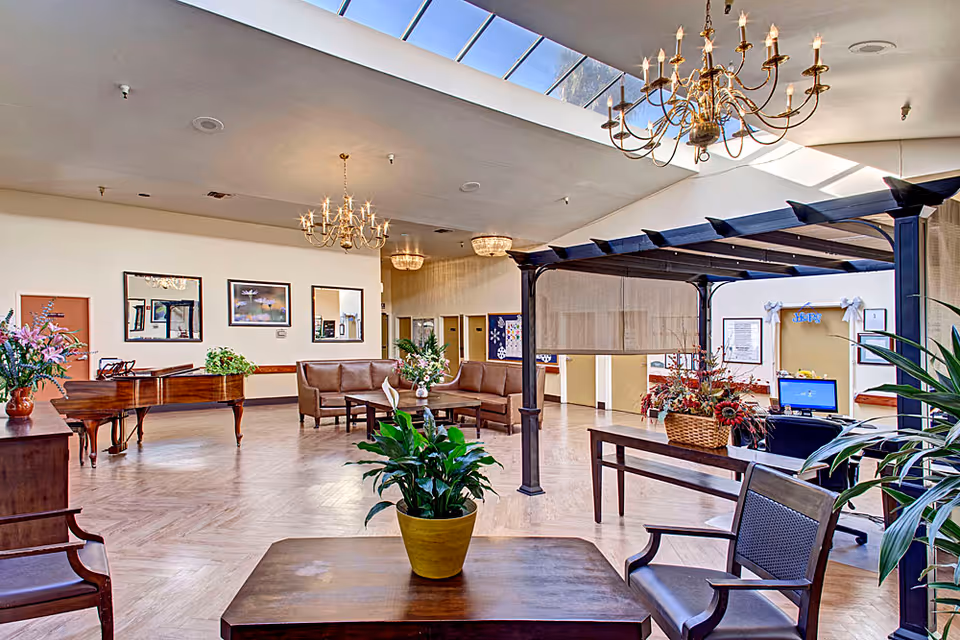 A spacious and well-lit common area in a senior living facility with wooden flooring, a grand piano, several brown leather sofas arranged around a coffee table with a flower vase, and a pergola structure with a table underneath decorated with a basket of flowers. The ceiling has chandeliers and a large skylight allowing natural light to fill the room. There are framed pictures on the walls and plants placed around the space.