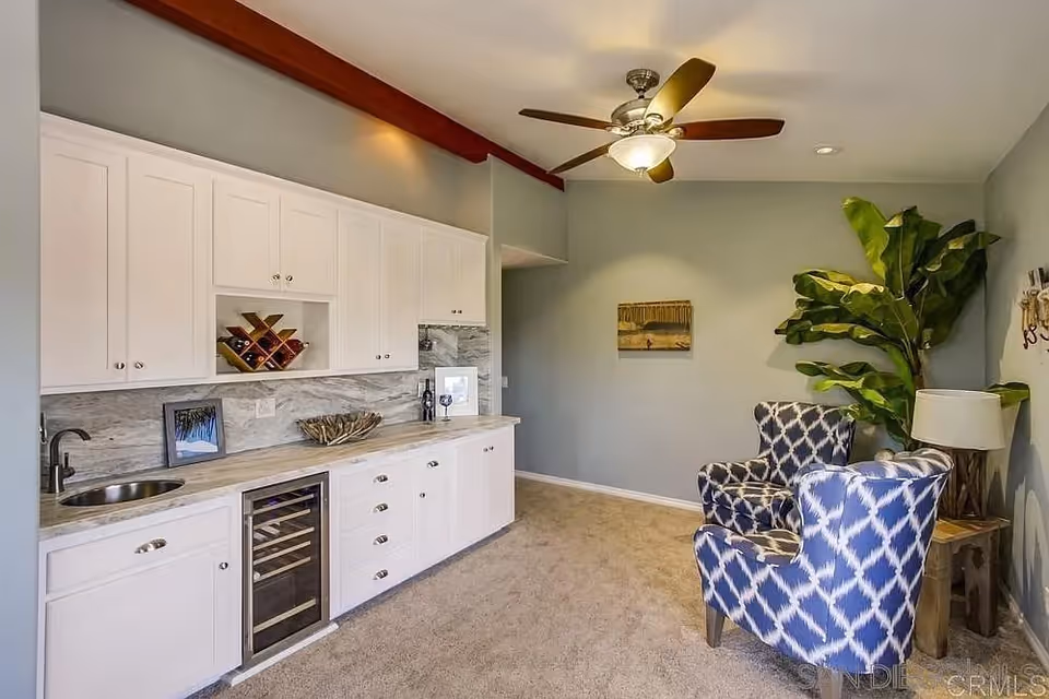 Bright interior sitting area with a built-in kitchenette, two patterned armchairs, a ceiling fan, and a large potted plant.