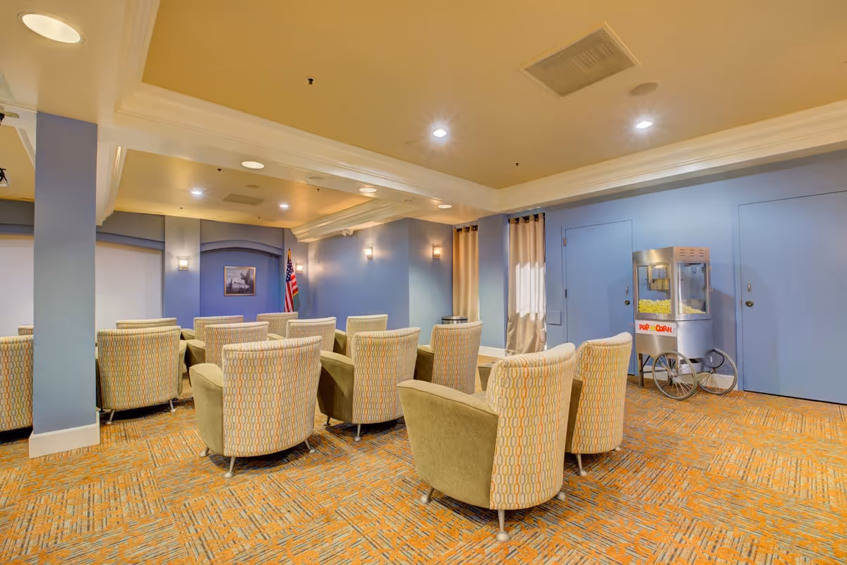 A cozy room with multiple upholstered armchairs arranged in rows facing a wall with an American flag and a framed picture. The room has blue walls, a popcorn machine on wheels, and a patterned carpet with orange and beige tones. The ceiling features recessed lighting and crown molding.